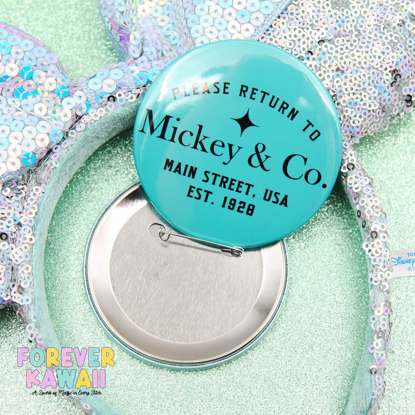 A turquoise pin badge with text, placed on a sequined background, with a silver pin attached to it.