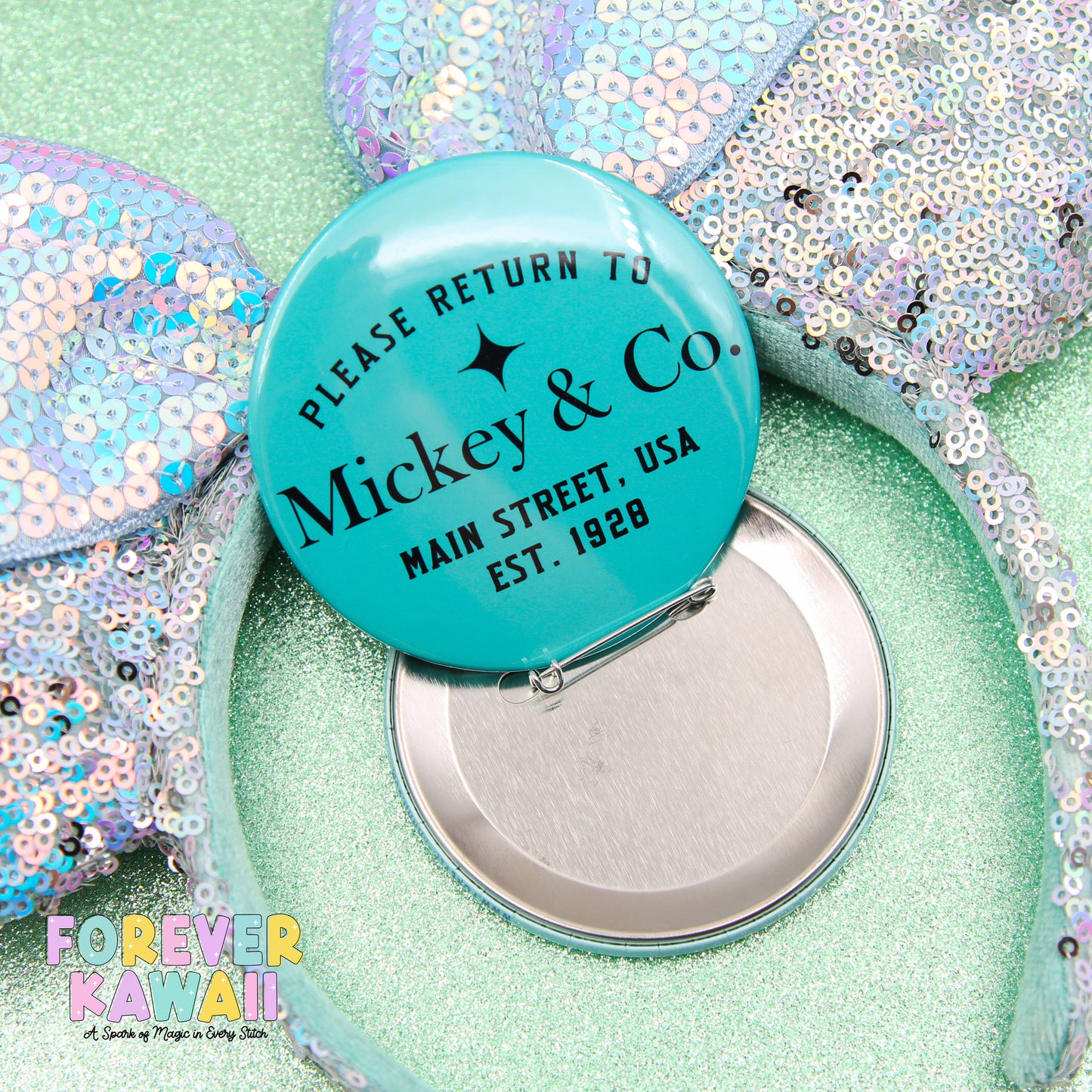 A turquoise Mickey & Co. pin with a silver button attached, placed on a glittery surface.
