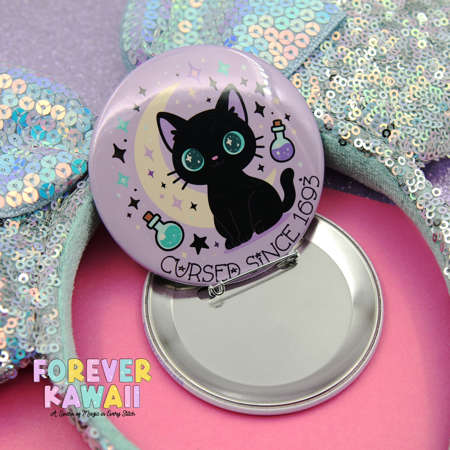 A round pin with a black cat design and a purple background, surrounded by stars and a crescent moon. The pin has a silver clasp and is placed on a pink surface.