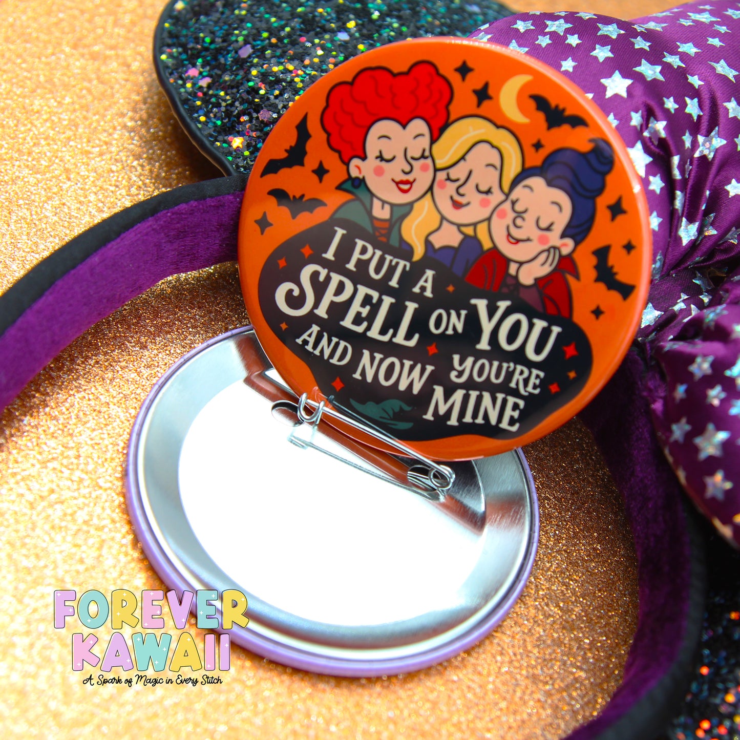 a round pin with a playful Halloween-themed design, including three cartoon girls with spooky hairstyles and a witch. The pin has a message that reads "I put a spell on you and now you're mine".