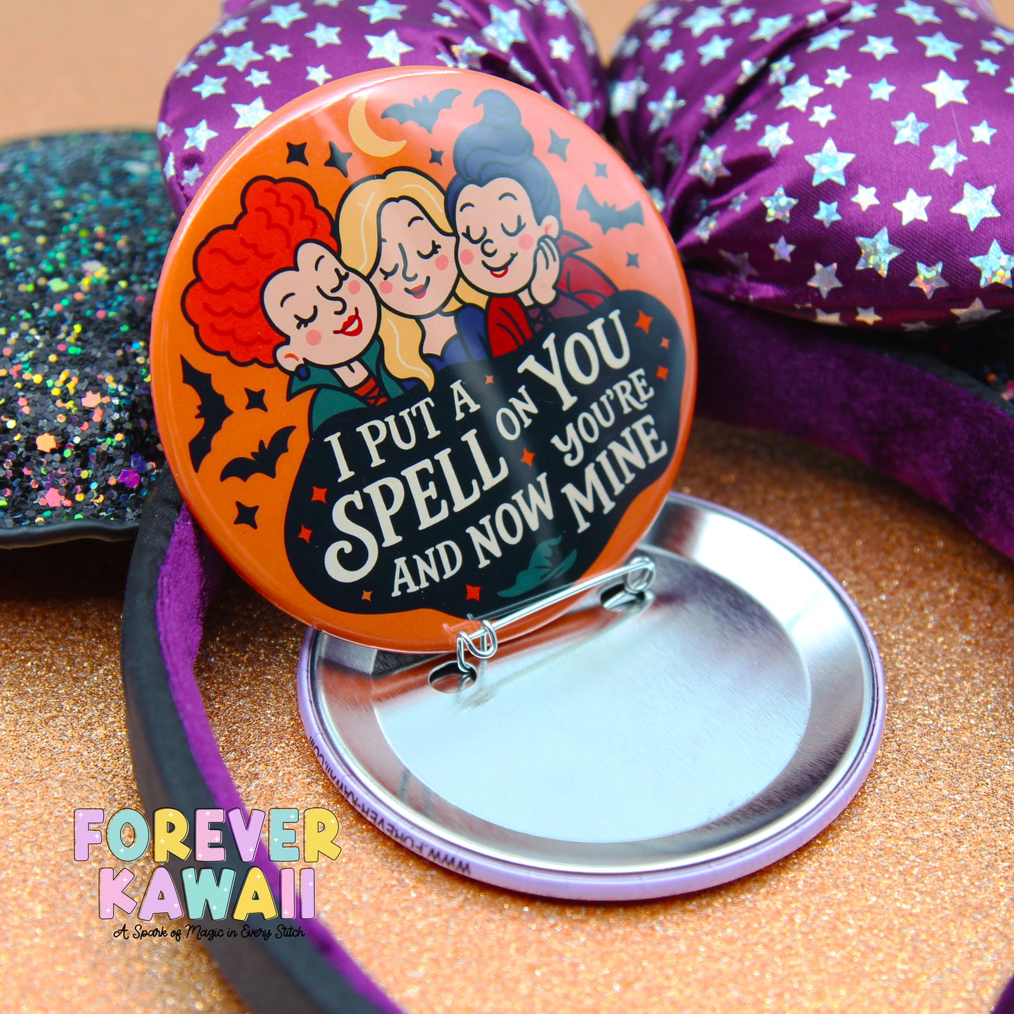 a round pin with a cartoon illustration of three people, along with text that says "I put a spell on you and now you're mine". The pin is placed on a purple and gold glittery surface, and there is a purple bow visible in the background.