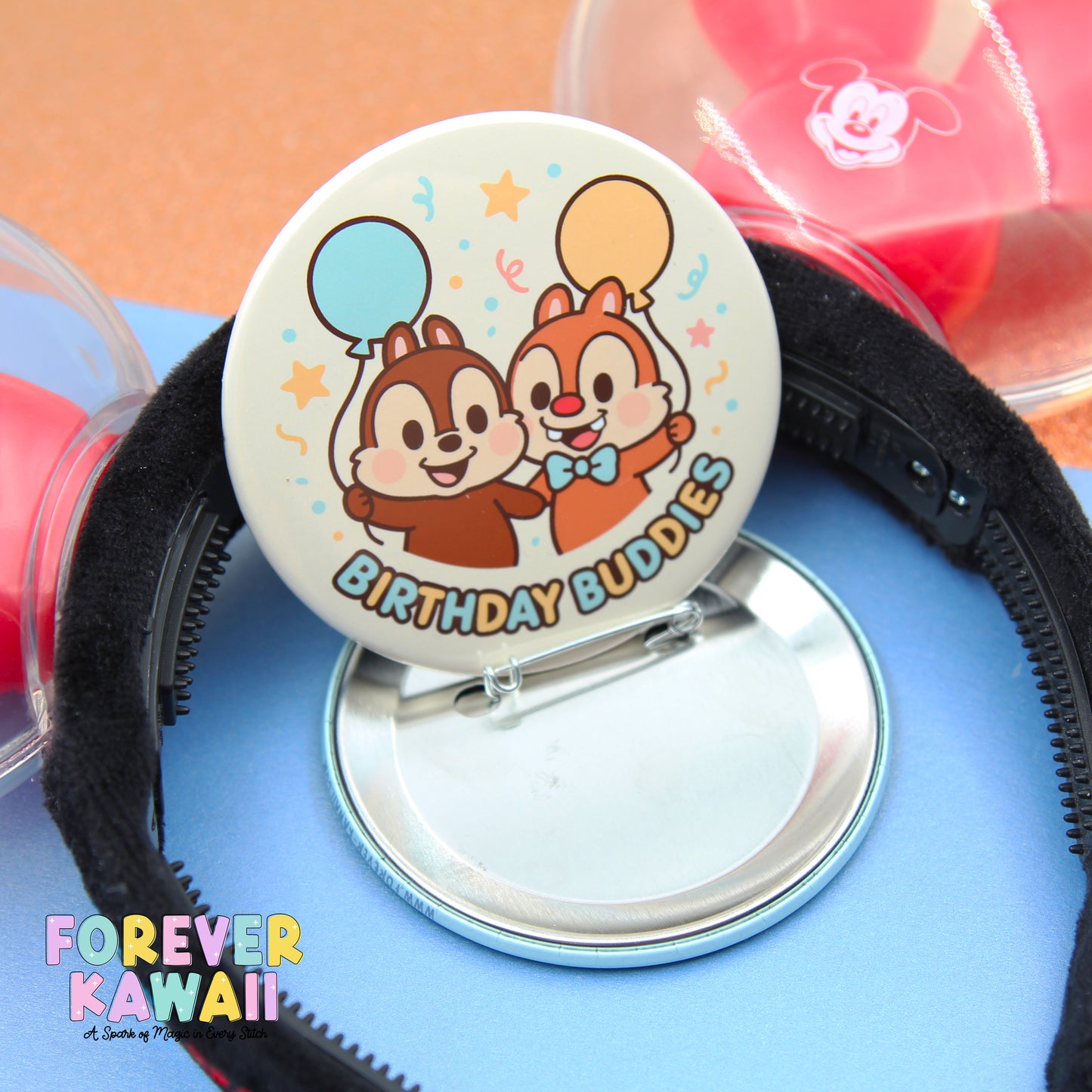 A round pin with a cartoon chip and two balloons, labeled "Birthday Buddies", is placed on a blue surface.