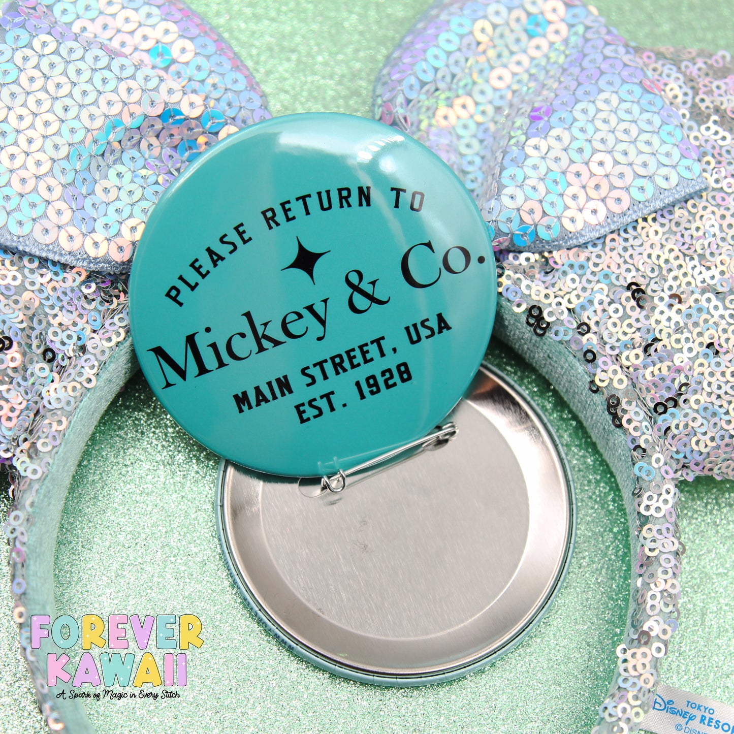 a turquoise pin with the text "Please Return to Mickey & Co. Main Street, USA est. 1928" on it, along with a silver pin lying on its side. The pin is surrounded by sequin-covered items, creating a colorful and playful scene.