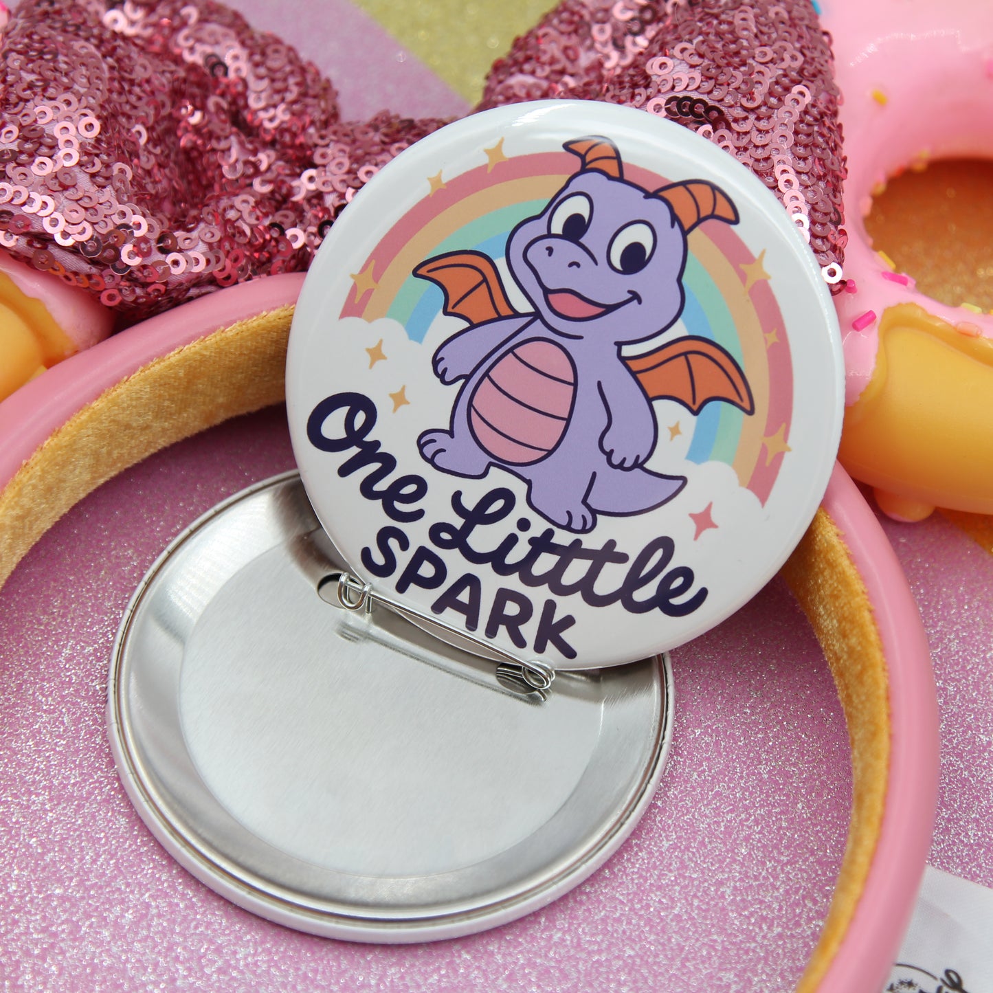A round pin with a cartoon dragon on it, surrounded by a pink sequin background.
