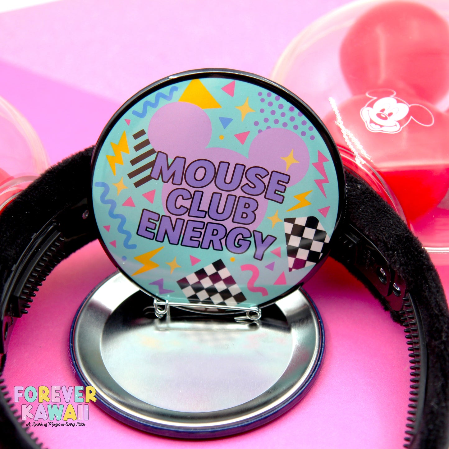 A round button with a colorful, retro-style design, featuring the text "MOUSE CLUB ENERGY" and various geometric shapes and patterns. The button is placed on a stand, and there are balloons visible in the background.