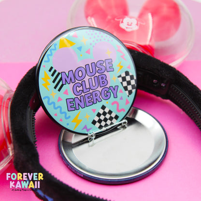 A colorful pin badge with the text "MOUSE CLUB ENERGY" on it, along with a playful design featuring a mouse and checkered patterns. The badge is placed on a pink surface, and there is a black headband visible in the background.