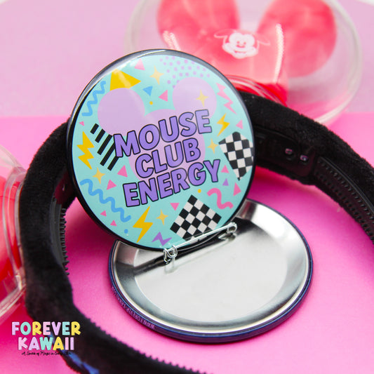 A colorful pin badge with the text "MOUSE CLUB ENERGY" on it, along with a playful design featuring a mouse and checkered patterns. The badge is placed on a pink surface, and there is a black headband visible in the background.