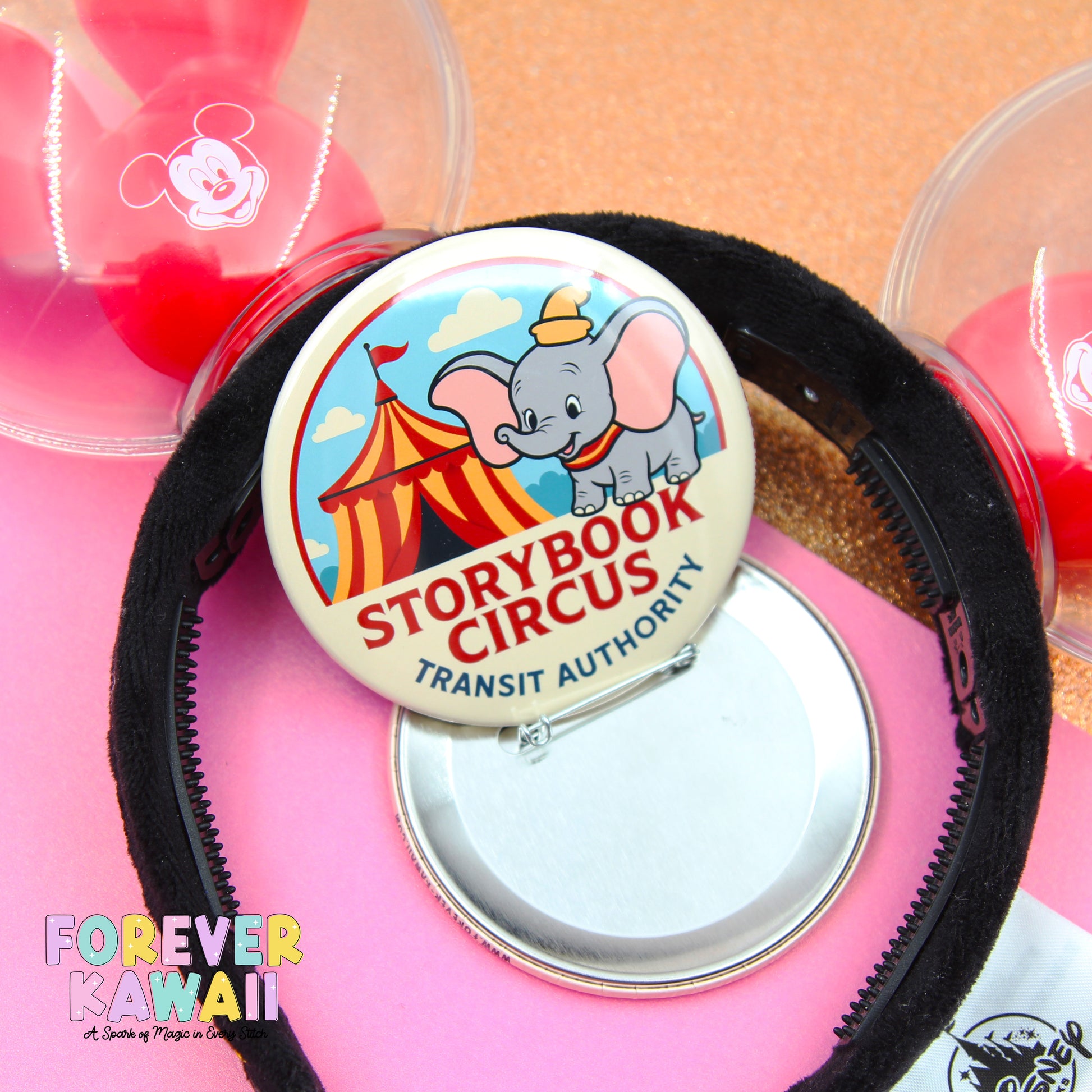 a pink background with a black headband, a round pin, and a clear container with a Mickey Mouse logo.