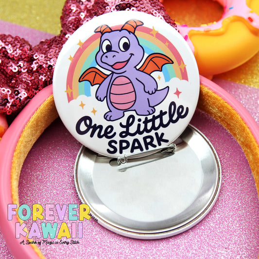 A pin with a dragon design and the text "One Little Spark" on it, surrounded by a pink sequin background and a pink headband.