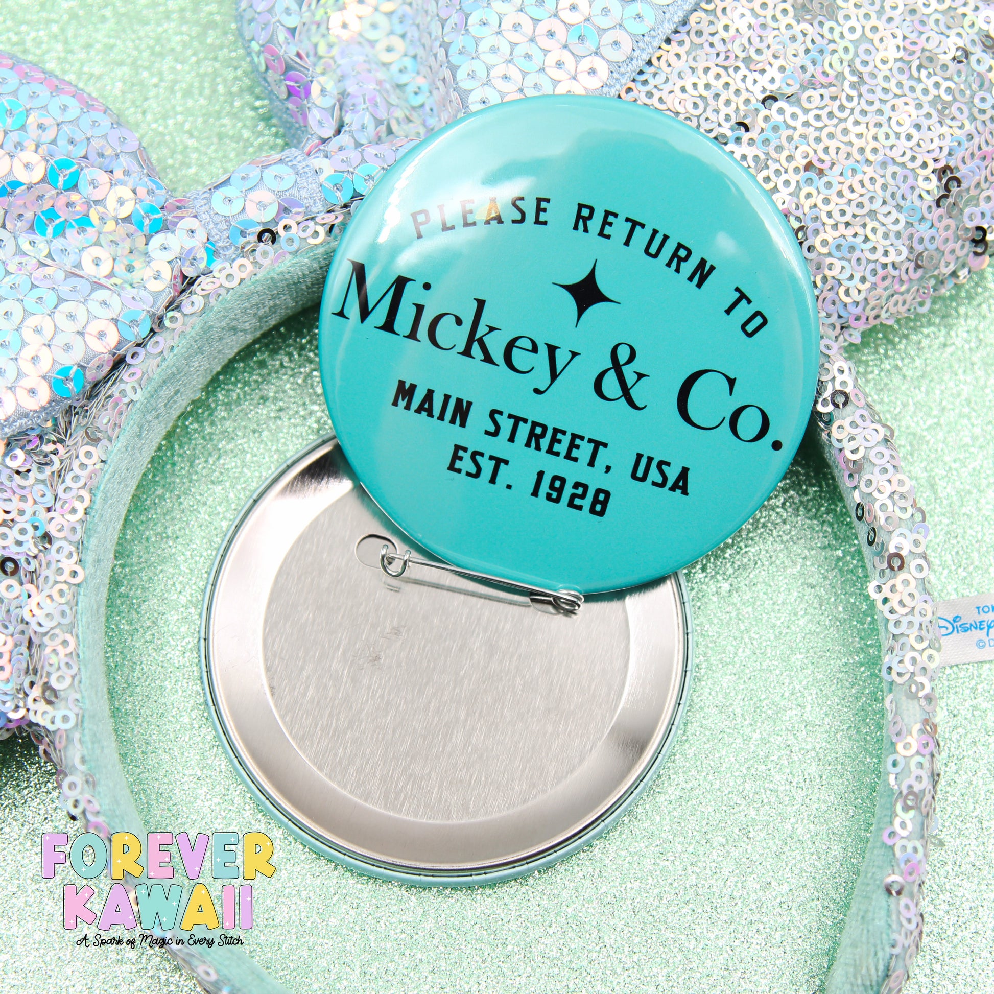 A turquoise pin badge with text, placed on a sequined background, with a silver pin attached to it.