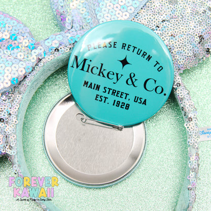 A turquoise pin badge with text, placed on a sequined background, with a silver pin attached to it.