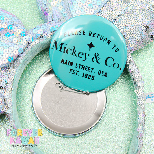 A turquoise pin badge with text, placed on a sequined background, with a silver pin attached to it.
