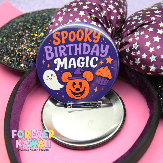 A round pin with a spooky birthday message, featuring a ghost, pumpkin, and cupcakes, is displayed on a pink surface.