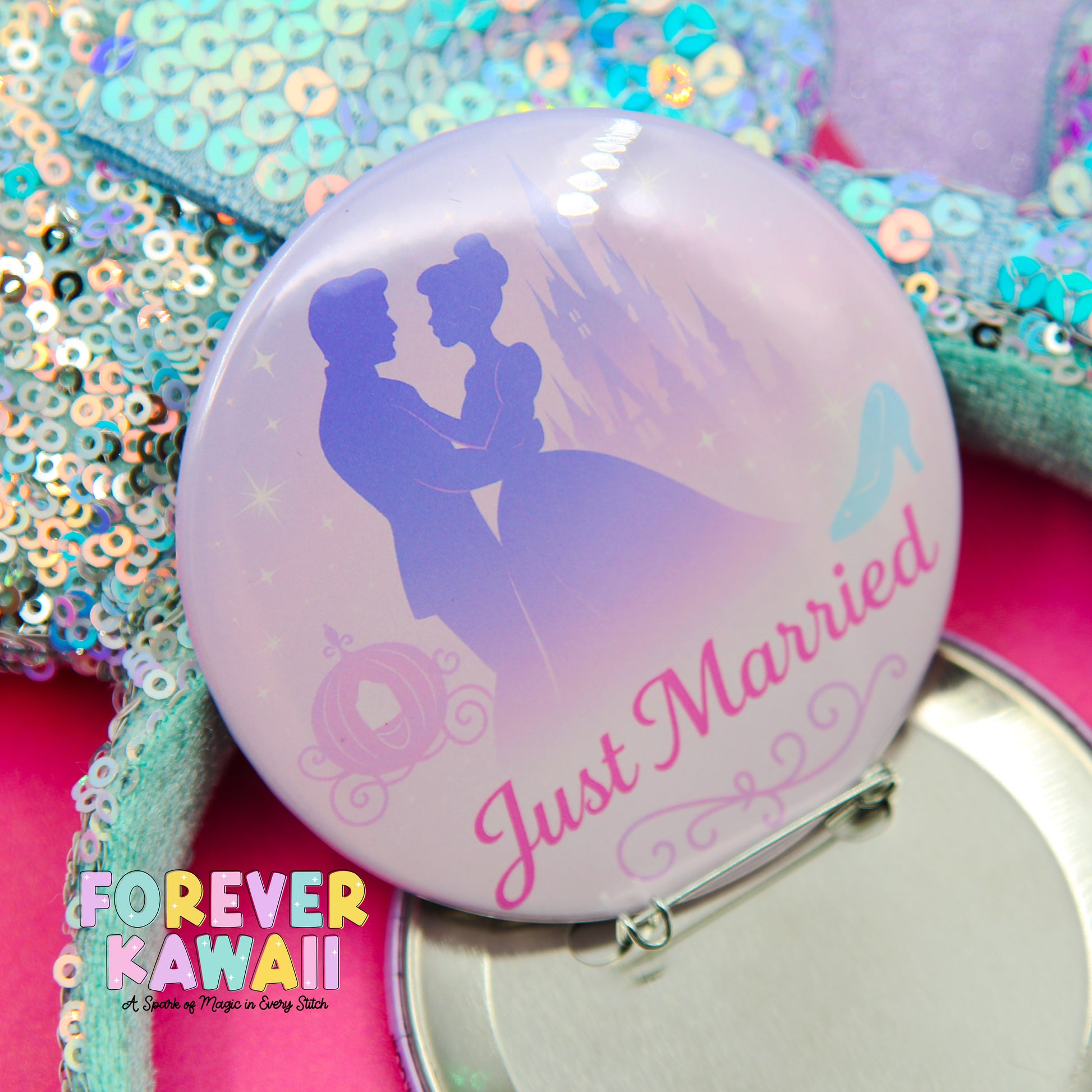 A round pin with a silhouette of a couple embracing, along with the text "Just Married" and a castle in the background. The pin is placed on a pink surface, surrounded by sequins and other decorative elements.