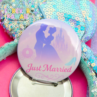 A button with a silhouette of a couple embracing, along with a castle and a carriage, and the text "Just Married" printed on it. The button is placed on a pink surface, and there is a sequined item in the background.