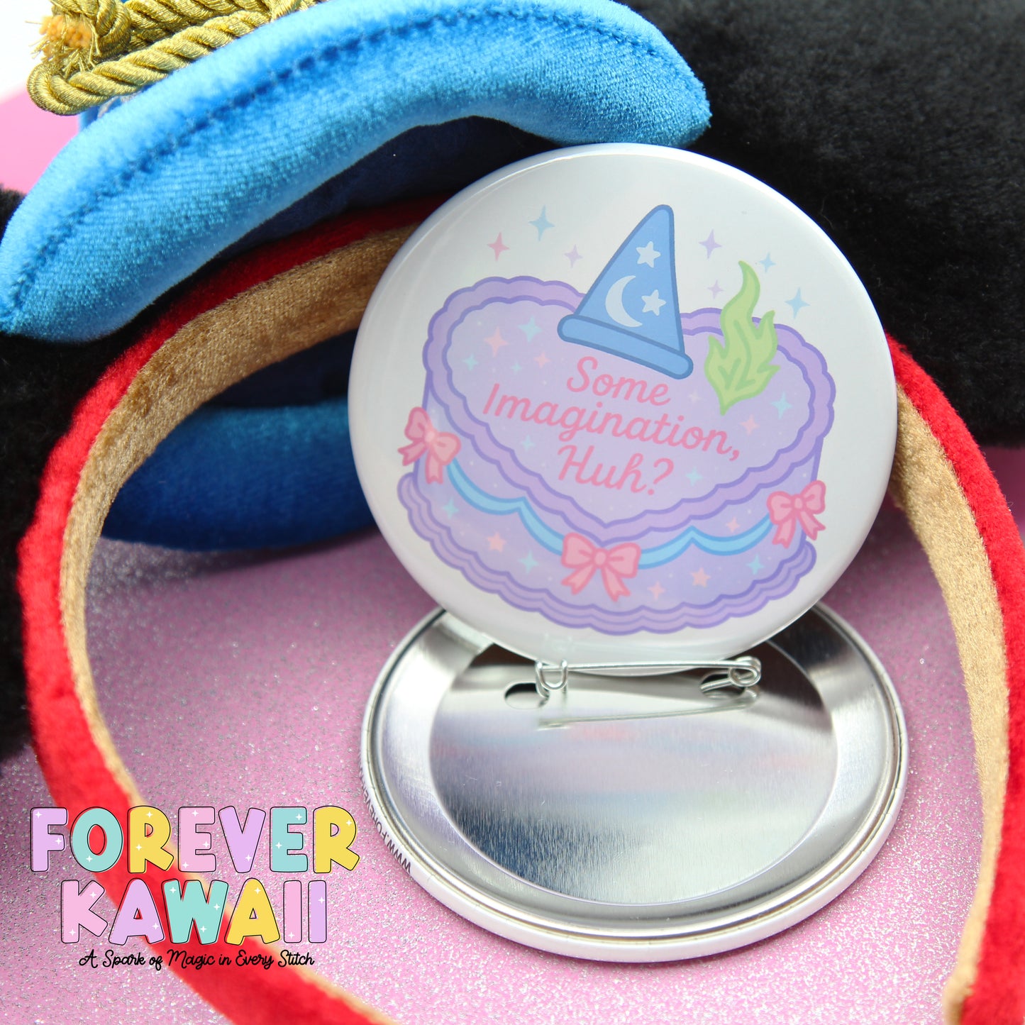 A round pin with a whimsical design, featuring a wizard hat and a cake with stars and flowers, is placed on a pink surface.