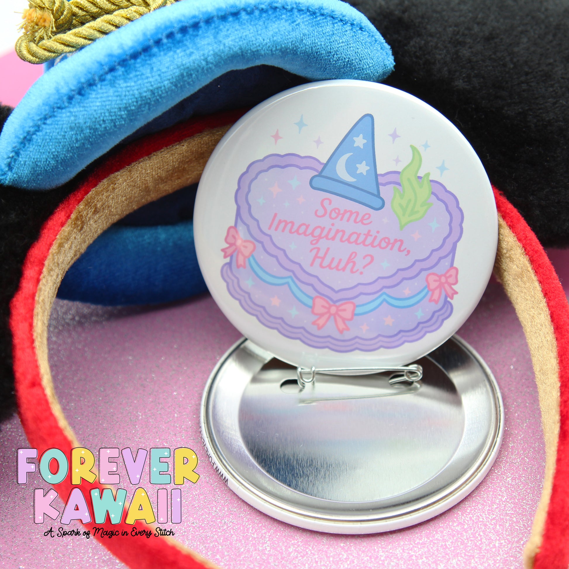 A round pin with a whimsical design, featuring a wizard hat and a cake with stars and flowers, is placed on a pink surface.