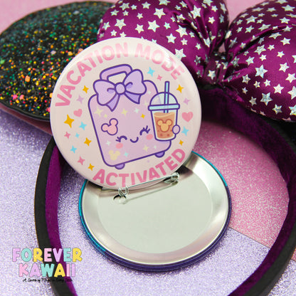 a round pin with a cartoon character and the text "VACATION MODE ACTIVATED" on it, placed on a reflective surface.