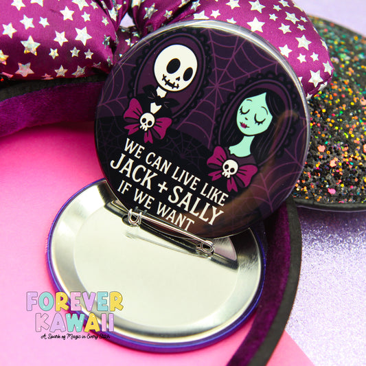 A round pin with a cartoon illustration of a skeleton and a green-skinned girl, along with the text "WE CAN LIVE LIKE JACK + SALLY IF WE WANT" printed on it. The pin is placed on a pink surface, with a purple bow and glittery objects in the background.