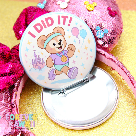 A round pin badge with a cartoon bear and the text "I DID IT!" printed on it, surrounded by sequin decorations and a pink sequin headband.