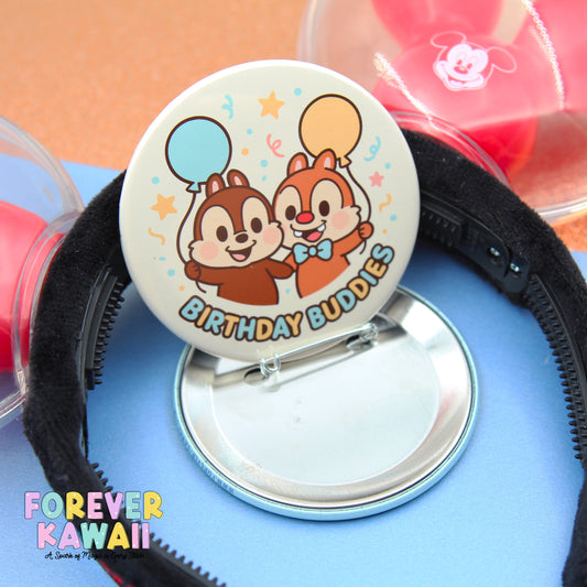 A round pin with a cartoon chip and two balloons, labeled "Birthday Buddies", is placed on a blue surface.
