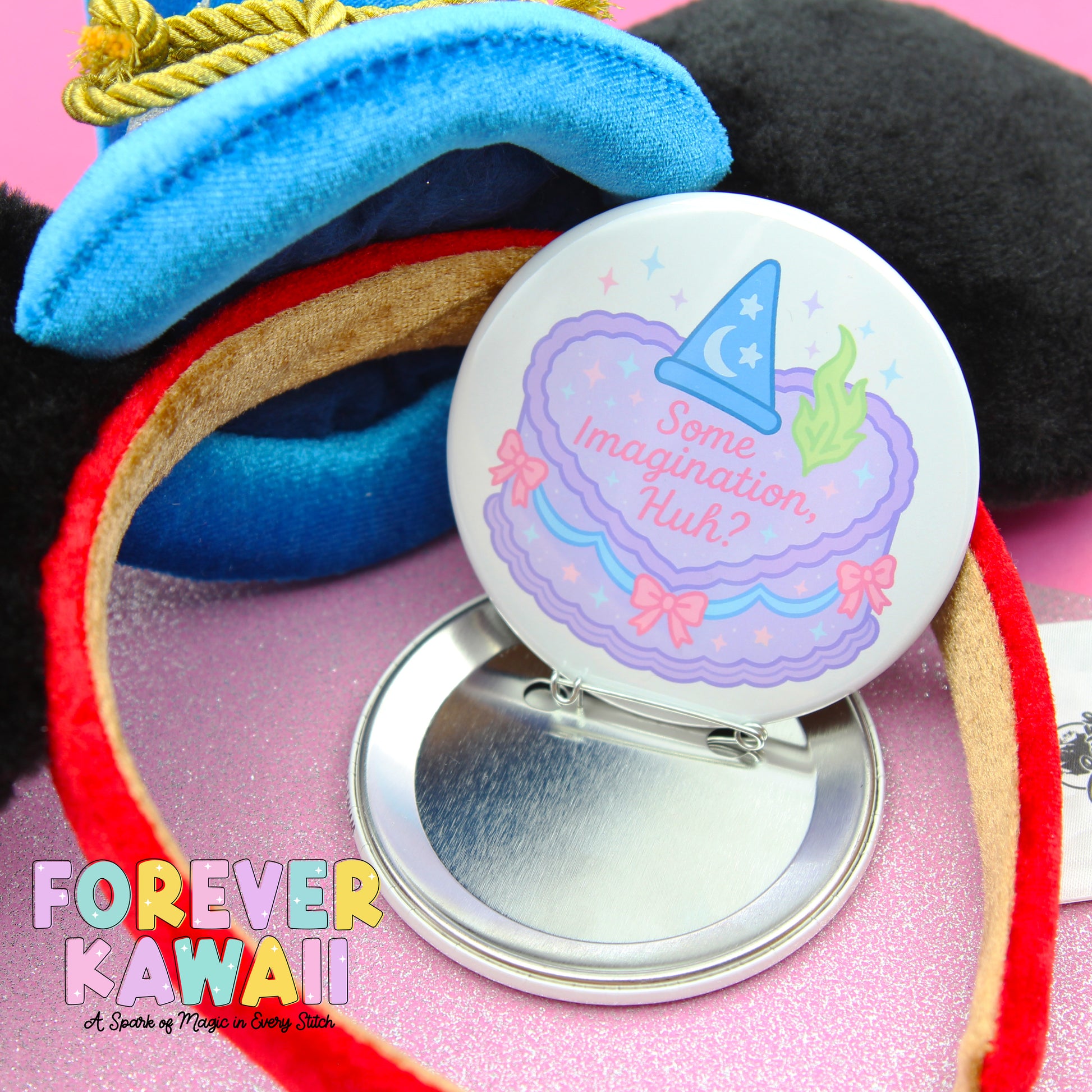 A round pin with a whimsical design, including a wizard hat and a cake with a message, is placed on a pink surface.
