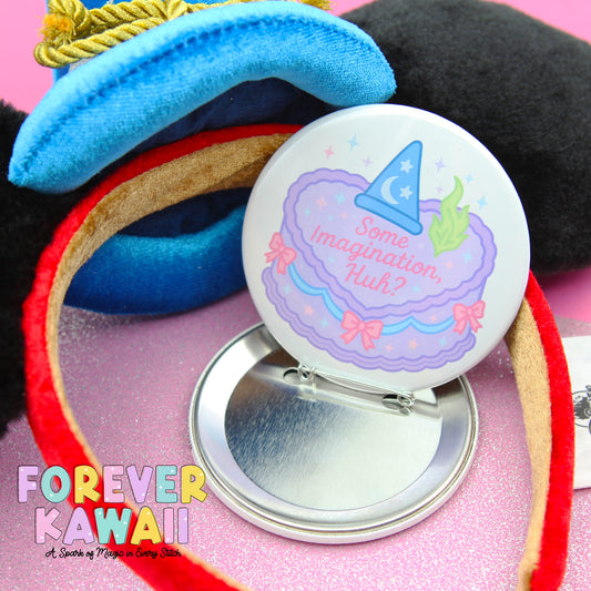 A round pin with a whimsical design, including a wizard hat and a cake with a message, is placed on a pink surface.