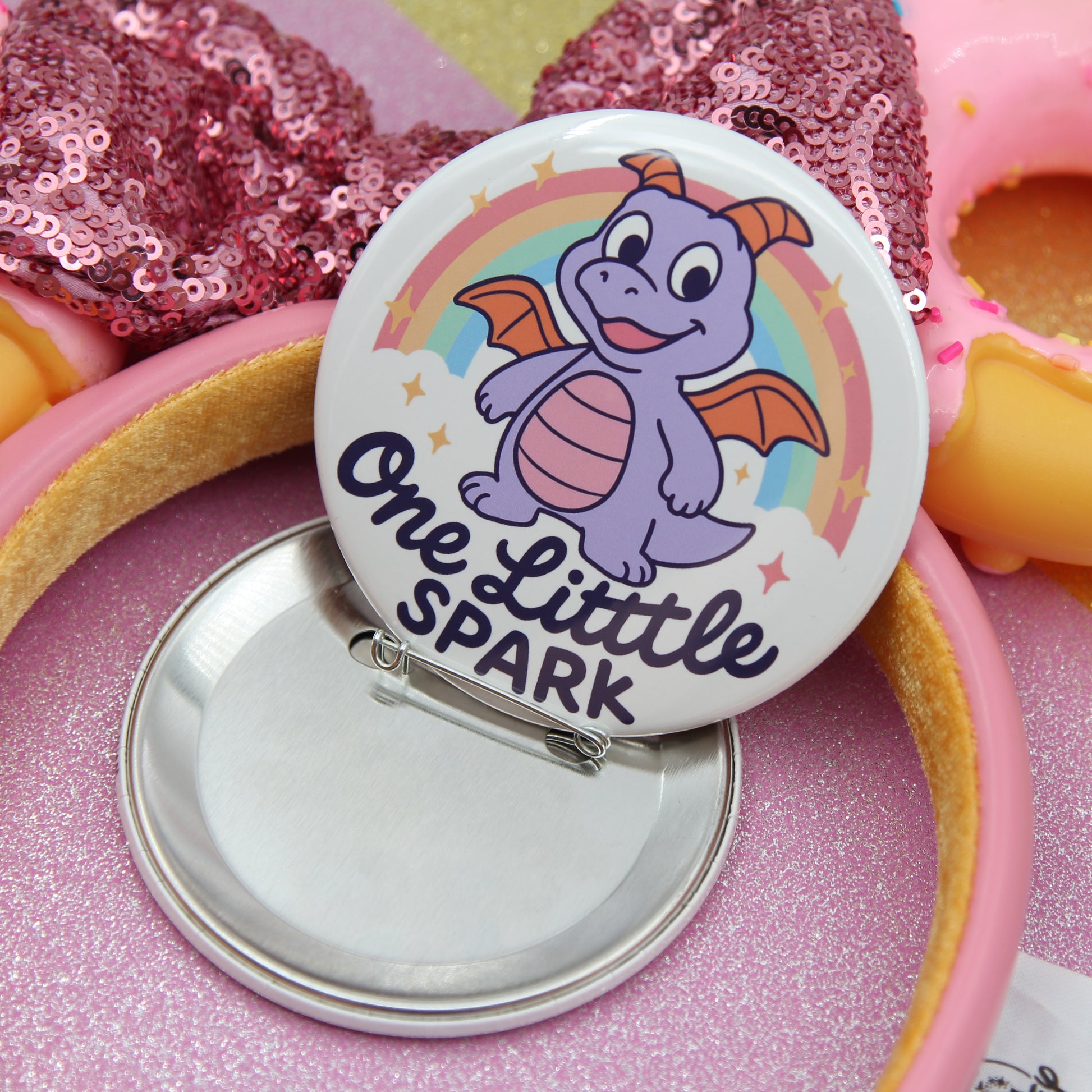 A round pin with a cartoon dragon on it, surrounded by a pink sequin background.