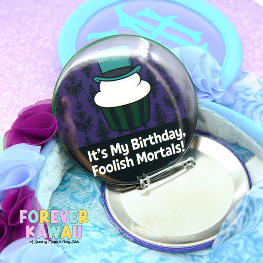 A round badge with a cartoon cupcake on it, surrounded by a blue and purple background. The badge has text that says "It's My Birthday, Foolish Mortals!"