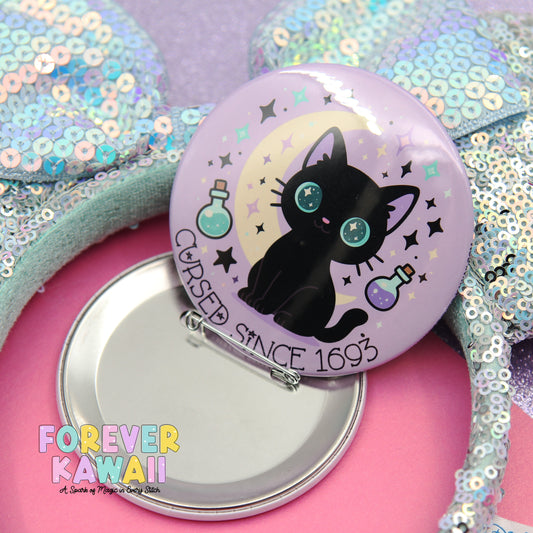 A round pin with a black cat design and stars on a purple background, along with a silver button and a pink background.