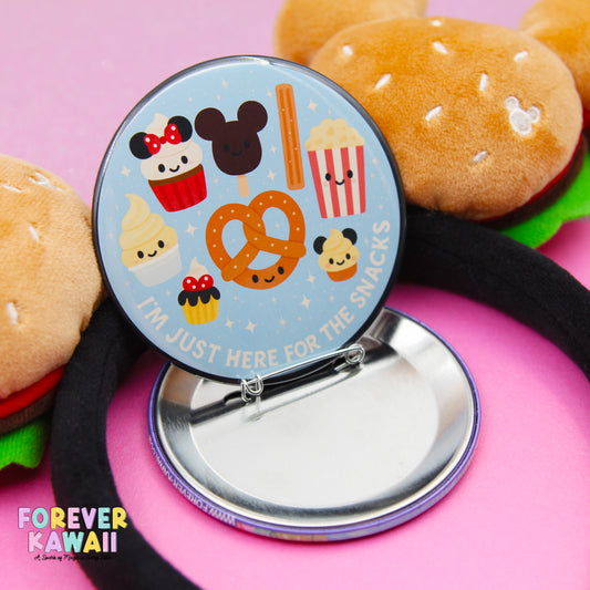 A round button with a playful design featuring various cartoon characters and snacks, such as a pretzel, cupcake, and popcorn, along with the text "I'M JUST HERE FOR THE SNACKS".