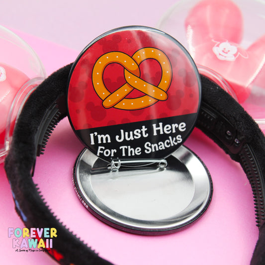 A red button with a yellow pretzel and the text "I'm Just Here For The Snacks" is shown, along with a black hairband.